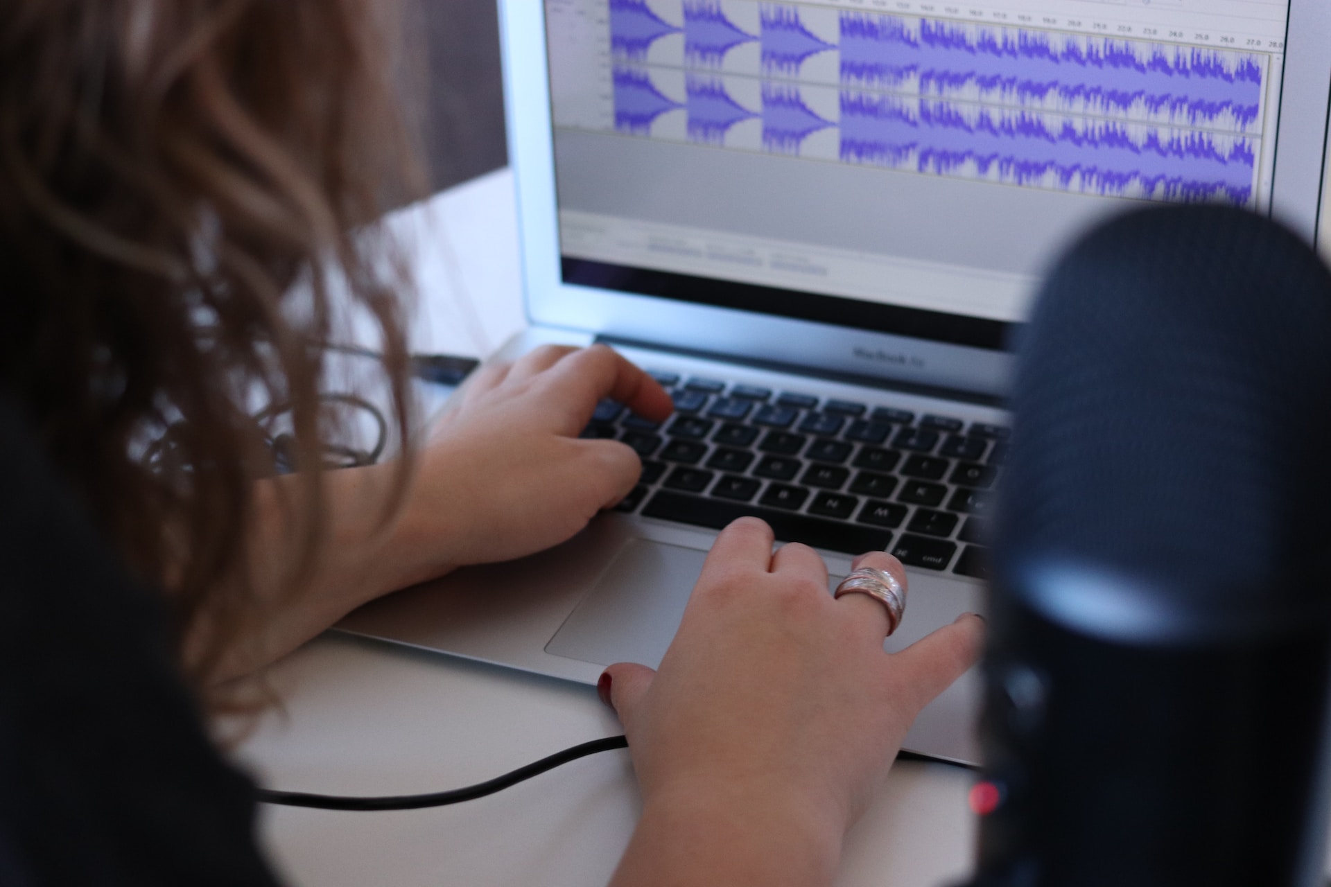 A person with long hair and rings on fingers at a laptop with some podcast editing software on the screen and a podcast microphone in shot